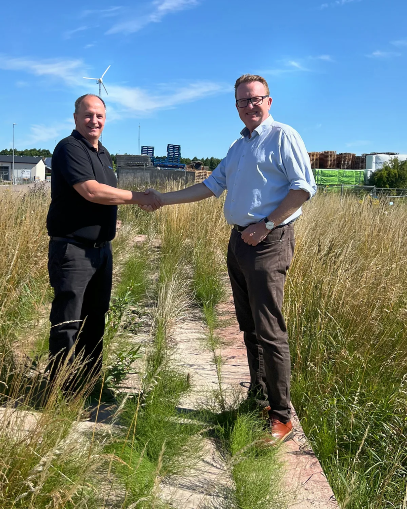 Johan Wester and Anders Malm shake hands on the plot at Kontoristgatan 2, with GLF’s pallet and timber storage area in the background.