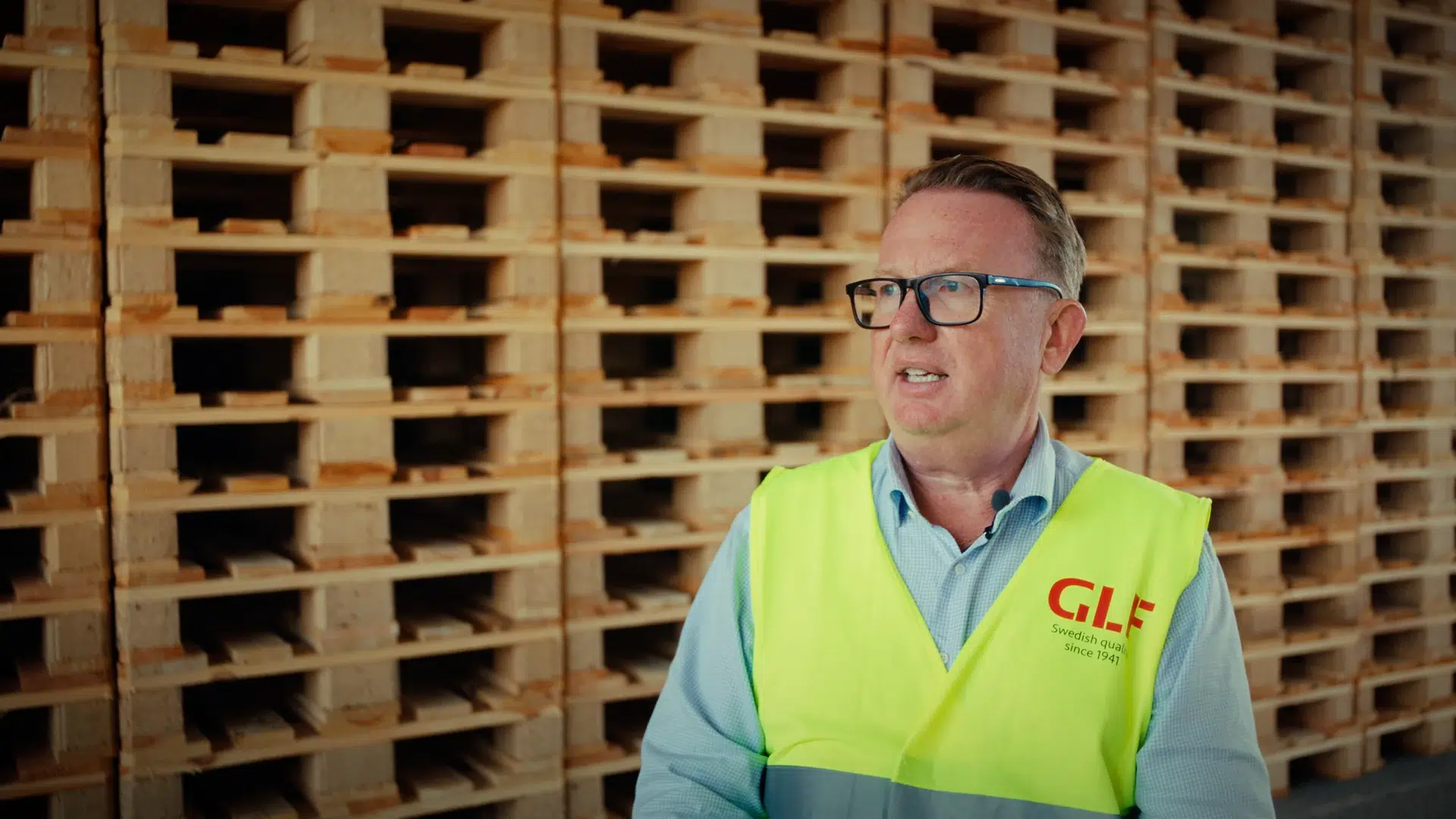 Une personne portant des lunettes et un gilet GLF jaune devant un mur de palettes en bois empilées.