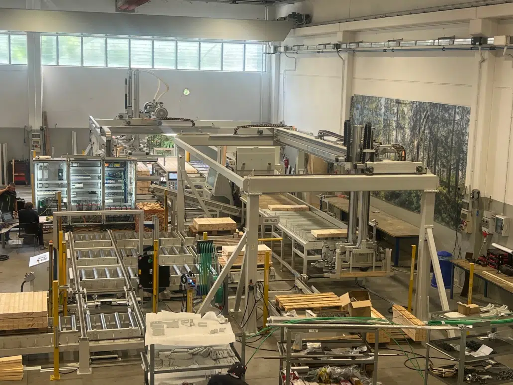Vue d’ensemble de la ligne de tronçonnage, avec ses convoyeurs à rouleaux, ses portiques et des paquets de bois dans le hall de l’usine.